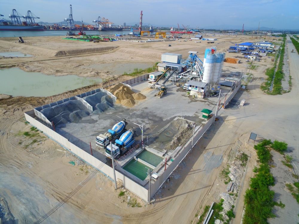 An aerial view of an industrial concrete batching plant with blue concrete mixer trucks, aggregate piles, and construction machinery, near a port with cranes and cargo containers in the background.